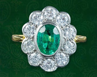 May include: An oval emerald gemstone ring with a halo of round-cut diamonds. The ring features a gold band and a silver setting around the emerald and diamonds. The ring is set against a dark green background.