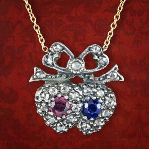Op de afbeelding: Een vintage hangerketting met twee hartvormige hangers, elk bezet met kleine, heldere edelstenen. Eén hart heeft een centrale roze edelsteen, de andere een blauwe. De ketting heeft een gouden ketting.