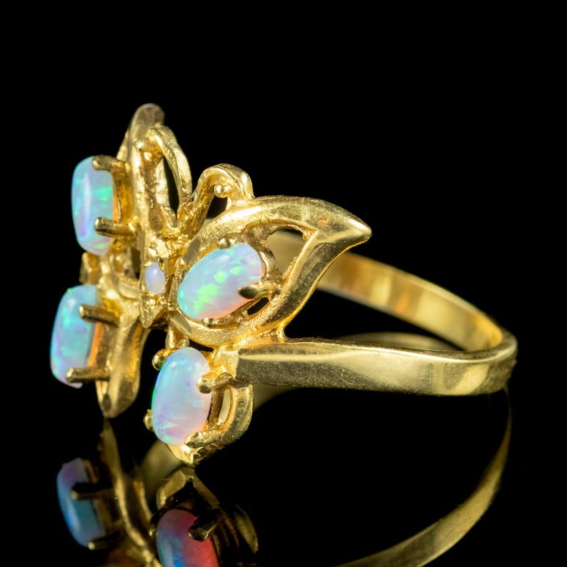 May include: A gold ring featuring a butterfly-shaped design set with five oval-cut opals. The metal band is smooth and polished, while the butterfly wings are detailed with prong settings that secure each iridescent white opal in place.