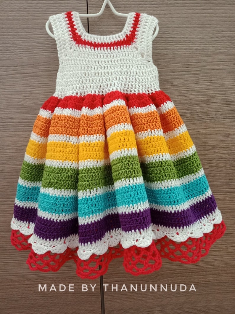 May include: A rainbow striped crochet dress with a white crochet bodice and a white crochet lace trim. The dress has a red crochet band at the neckline and a red crochet band at the hem.
