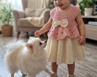 May include: A crochet dress featuring a pink bodice with a scalloped overlay and a cream-colored skirt, accented by a central cream bow. The text PDF PATTERN and Made by Thanunnuda are visible. A small white dog stands beside the dress.