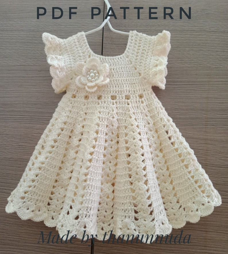 May include: A crocheted baby dress in a cream color with a flower embellishment. The dress has a scalloped hem and short sleeves. The dress is made with a delicate crochet pattern.