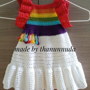 May include: A white crochet dress with a rainbow striped bodice and red crocheted sleeves. The dress has a ruffled hem and a purple tassel with colourful beads.