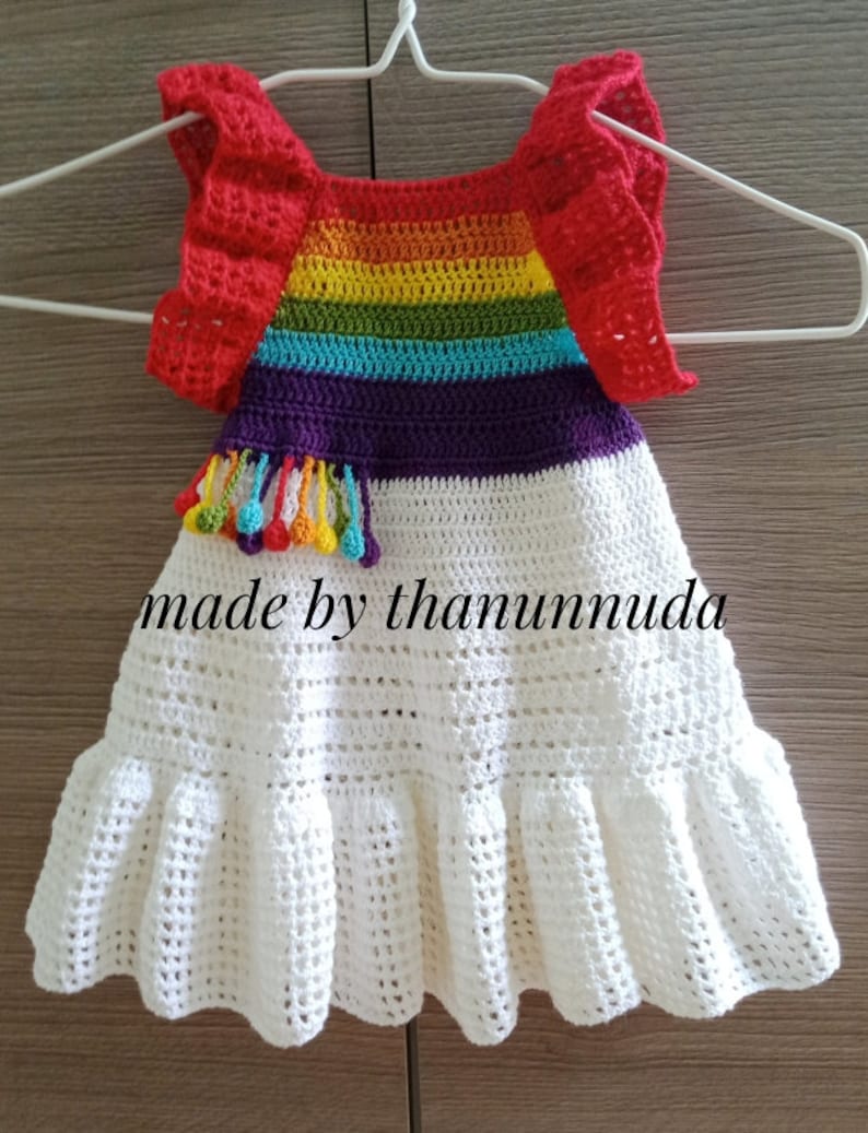 May include: A white crochet dress with a rainbow striped bodice and red crocheted sleeves. The dress has a ruffled hem and a purple tassel with colourful beads.