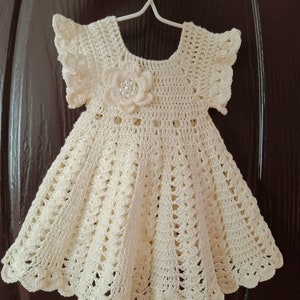 May include: A crocheted baby dress with a white flower embellishment. The dress has a scalloped hem and short sleeves. The text "PDF PATTERN" and "MADE BY THANUNNUDA" is visible in the image.