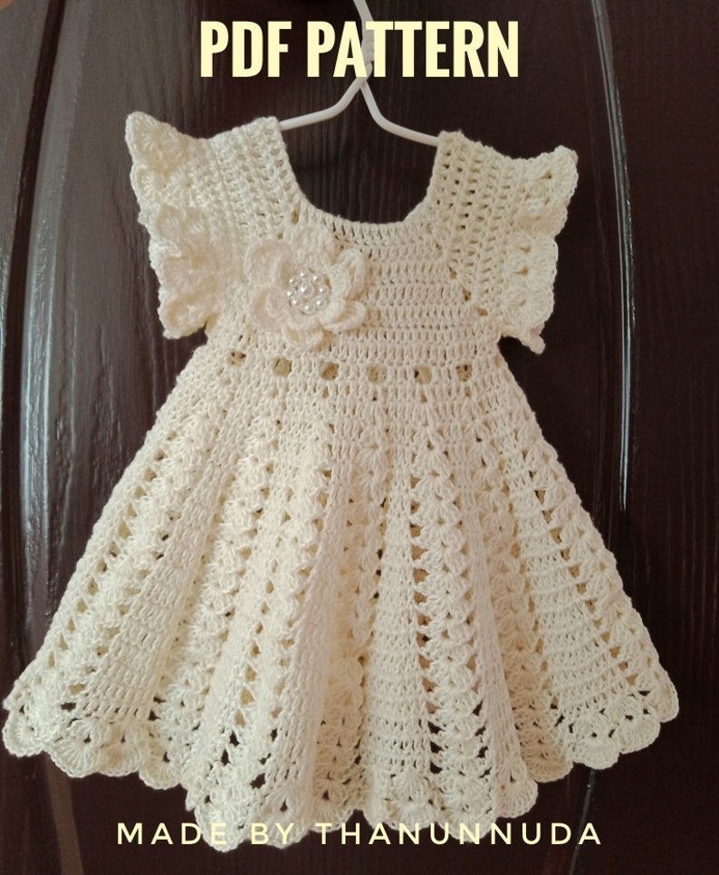 May include: A crocheted baby dress with a white flower embellishment. The dress has a scalloped hem and short sleeves. The text "PDF PATTERN" and "MADE BY THANUNNUDA" is visible in the image.