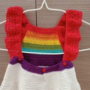 May include: A crocheted baby romper with a rainbow pattern on the bodice and red straps. The romper has a white and purple striped bottom with red buttons.