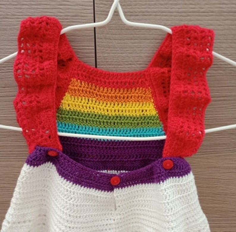 May include: A crocheted baby romper with a rainbow pattern on the bodice and red straps. The romper has a white and purple striped bottom with red buttons.