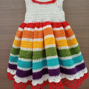 May include: A white crochet dress with rainbow stripes and a red crochet trim. The dress has a scalloped hem and is made with a fine yarn.