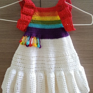 May include: A white crochet baby dress with a rainbow striped bodice and red ruffled sleeves. The dress has a white crocheted skirt with a ruffle at the hem. The bodice has a purple stripe and a rainbow tassel detail. The dress is made by thamunnuda.