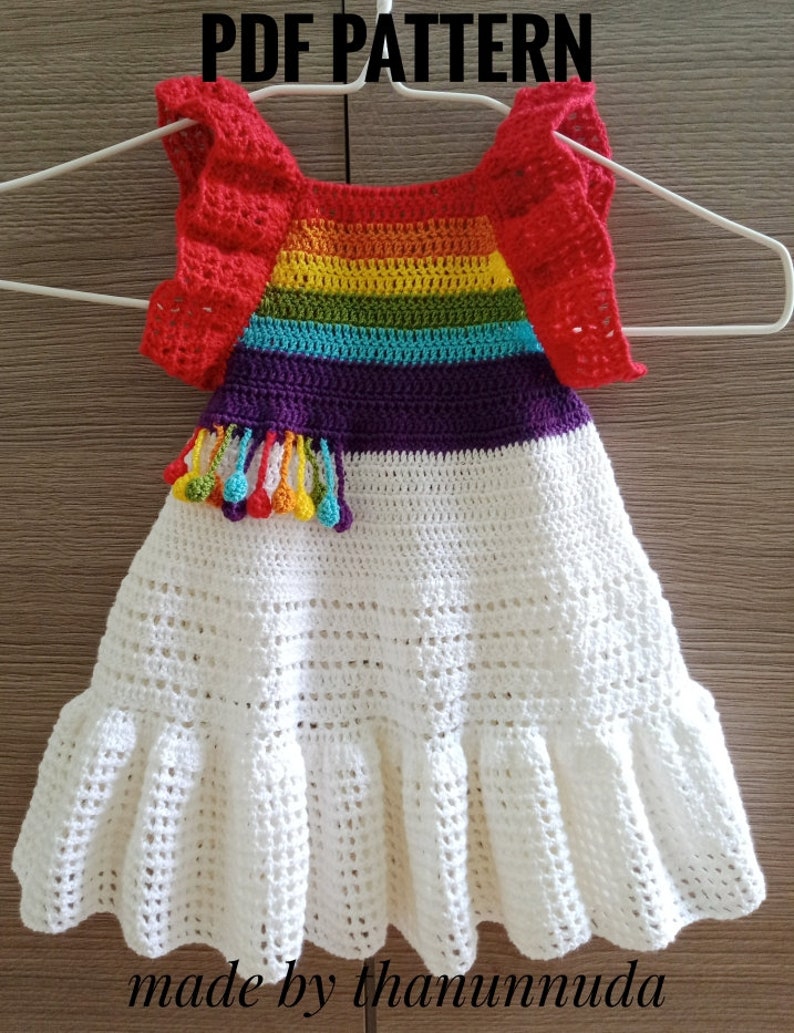 May include: A white crochet baby dress with a rainbow striped bodice and red ruffled sleeves. The dress has a white crocheted skirt with a ruffle at the hem. The bodice has a purple stripe and a rainbow tassel detail. The dress is made by thamunnuda.