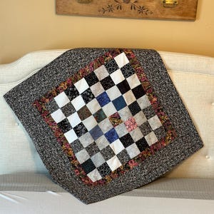 May include: A black and white checkered quilt with a floral border. The quilt is made of squares of different fabrics and is folded in half. It is sitting on a white bed.