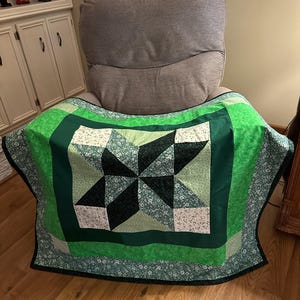May include: A green and black quilt with a star pattern. The quilt is folded and sitting on a chair.