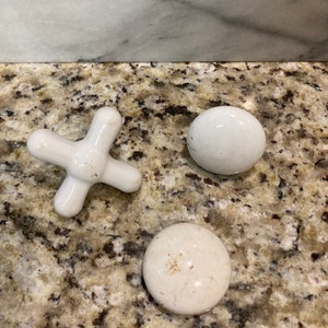 May include: Three white ceramic knobs, one shaped like an X and two round. The knobs are on a brown and white speckled surface.