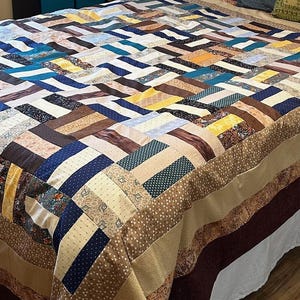 May include: A patchwork quilt with a geometric pattern in shades of brown, blue, beige, and yellow. The quilt is made of various fabrics with different textures and patterns.