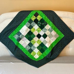 May include: A handmade quilt with a St. Patrick's Day theme. The quilt features a central diamond pattern of green, white, and black squares, framed by a bright green border and a dark green backing. The quilt is displayed on a white surface.
