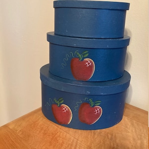 May include: Three blue nesting boxes with painted red apples. The largest box has two apples, the middle box has one apple, and the smallest box is plain.
