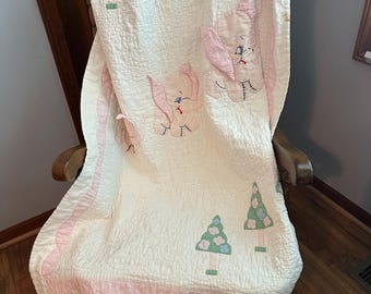 Puede incluir: Una colcha de bebé vintage con un fondo crema, con conejitos rosas y árboles verdes y azules aplicados. La colcha tiene un borde rosa festoneado y está sobre una mecedora de madera.