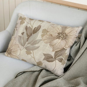 May include: A beige throw pillow with a floral pattern featuring brown and beige flowers and leaves. The pillow is on a light blue couch with a brown and white throw blanket.