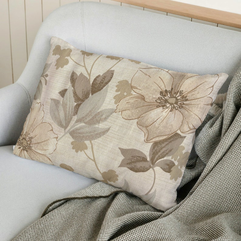 May include: A beige throw pillow with a floral pattern featuring brown and beige flowers and leaves. The pillow is on a light blue couch with a brown and white throw blanket.