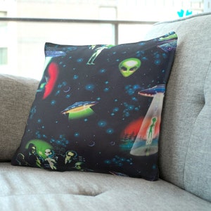 May include: A black and green throw pillow with a repeating pattern of aliens, UFOs, and stars. The pillow features a group of men in suits and a green alien in a beam of light.