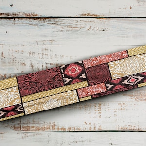 May include: A colourful fabric table runner with a patchwork design featuring geometric shapes and floral patterns in shades of red, gold, and white. The runner is laid diagonally on a whitewashed wooden surface.