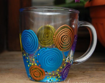 Pode incluir: Uma caneca de vidro transparente com alça, pintada à mão com redemoinhos e pontos coloridos em tons de azul, amarelo, laranja e roxo.
