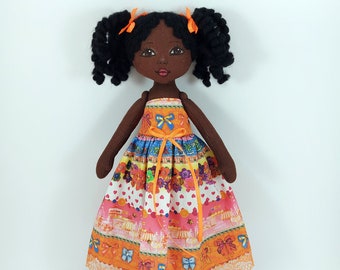 May include: A handmade doll with brown skin, black curly hair, and orange bows. She is wearing a colorful dress with a white background and orange trim.