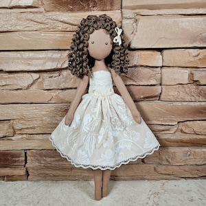 May include: A handmade doll with brown curly hair, wearing a white lace dress with a brown bodice and a small yellow bow in her hair.