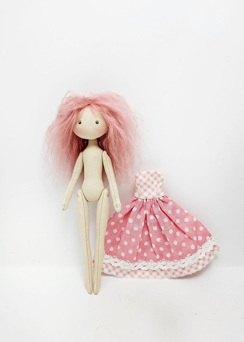 May include: A handmade doll with pink yarn hair and a pink and white polka dot dress. The doll has a simple, sewn-on face with black eyes and a pink mouth.