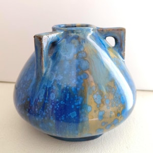 Può includere: Un vaso in ceramica con corpo arrotondato e due manici laterali. La superficie presenta uno smalto lucido con un motivo maculato nei toni del blu profondo, blu chiaro e marrone dorato, creando un effetto cristallino su tutto l'esterno.