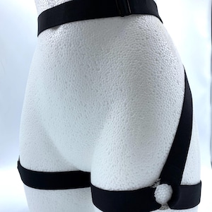 Pode incluir: Arnês de corpo elástico preto com cintas ajustáveis na cintura e na coxa. O arnês apresenta um detalhe de anel de metal nas cintas da coxa e um fecho de fivela na cintura. Exibido em um manequim branco.