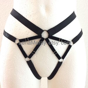 Puede incluir: Arnés de lencería negro con anillos de metal plateado. El arnés tiene un diseño geométrico con múltiples correas y anillos.