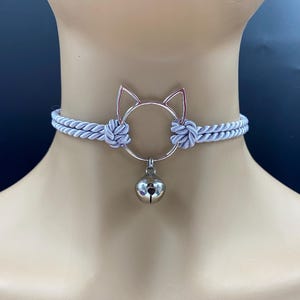 Puede incluir: Un collar gargantilla de cuerda gris claro con un colgante de plata en forma de gato y un pequeño dije de campana.