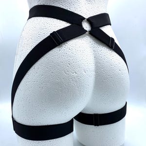 Può includere: Harness nero con cinghie regolabili e un dettaglio ad anello in metallo. L'imbracatura presenta una fascia per la coscia e un design a incrocio, esposta su un manichino bianco. La lingerie è realizzata in materiale elastico.