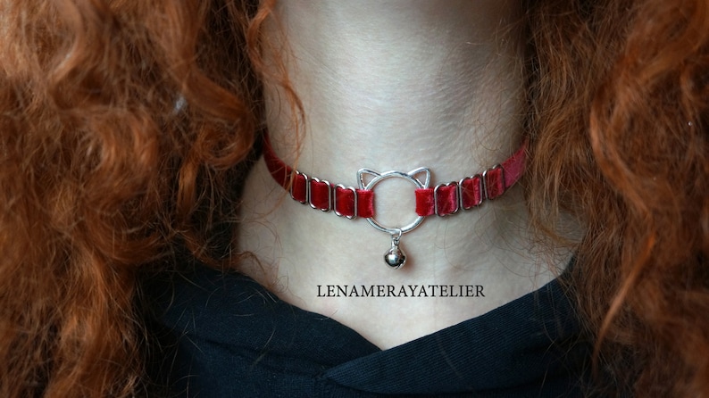 May include: A red velvet choker with a silver cat-shaped charm and a bell. The choker is adjustable and has a delicate, feminine look.