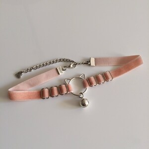 May include: A pink velvet choker necklace with a silver cat charm and a bell. The choker has a chain extender and a small heart charm.