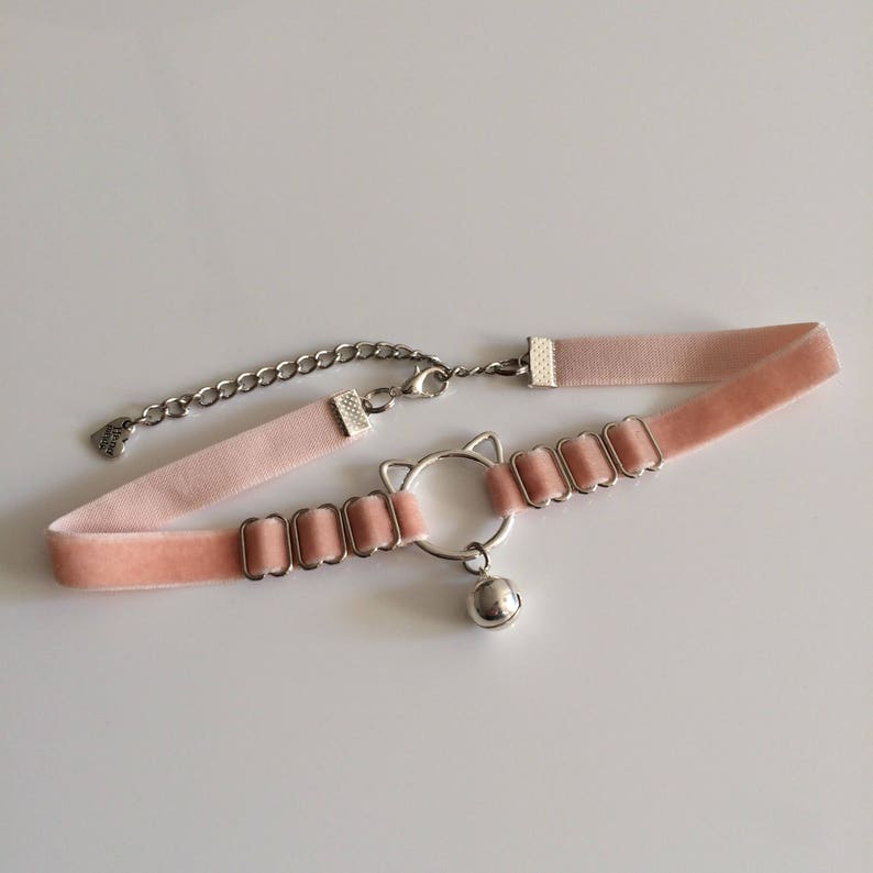May include: A pink velvet choker necklace with a silver cat charm and a bell. The choker has a chain extender and a small heart charm.