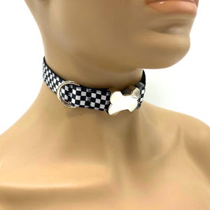 Könnte beinhalten: Ein schwarz-weiß kariertes Halsband mit einem silberfarbenen Metall-D-Ring und einem knochenförmigen Schnallenverschluss. Das Halsband besteht aus einem gewebten Stoff mit einem sich wiederholenden Quadratmuster und polierten Metallbeschlägen.