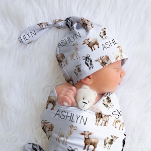May include: A white swaddle with a goat print and the name "ASHLYN" repeated throughout. A baby is wearing the swaddle and a matching hat. The baby is holding a stuffed animal.