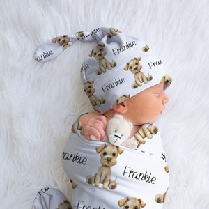 May include: A white swaddle with a pattern of brown puppies and the name 