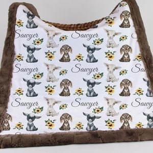 May include: White baby blanket with a brown faux fur border. The blanket features a repeating pattern of cute cartoon goats and sunflowers. The name "Sawyer" is printed multiple times on the blanket.
