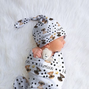 May include: A newborn baby swaddled in a white blanket and matching hat, both adorned with a repeating pattern of pug dog illustrations, paw prints, and the name "Harrison". The baby is nestled on a fluffy white surface.