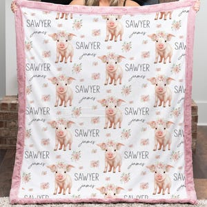 May include: A white baby blanket with a pink border features a repeating pattern of cartoon pigs wearing floral crowns and the name "Sawyer James" in cursive. The blanket is made of soft material.
