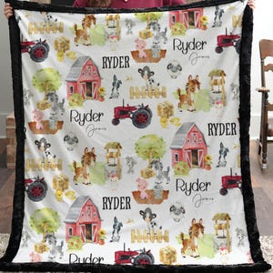 May include: A white blanket with a black border featuring a farm animal pattern. The design includes red barns, tractors, horses, pigs, and the name "Ryder" in black script. The blanket is held up by a person.