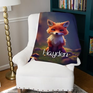 May include: A soft blanket with a cartoon fox design in orange and white, set against a blurred forest backdrop. The name "Hayden" is written in white script. The blanket is draped over a white armchair.