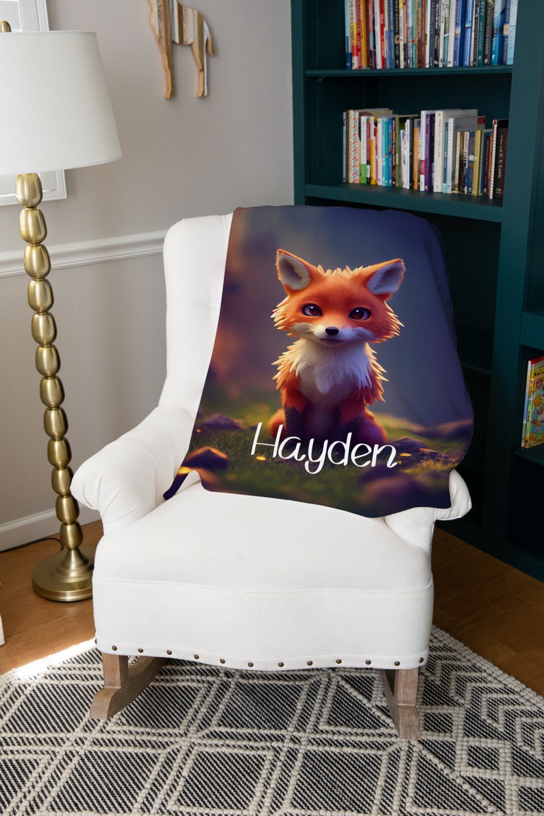May include: A soft blanket with a cartoon fox design in orange and white, set against a blurred forest backdrop. The name "Hayden" is written in white script. The blanket is draped over a white armchair.