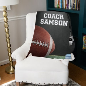 Pu&ograve; includere: Una coperta in pile nero e grigio con il testo "COACH SAMSON" in bianco. Un pallone da football marrone e bianco si trova nell'angolo in basso a sinistra della coperta.