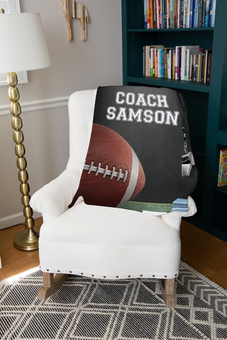 Pu&ograve; includere: Una coperta in pile nero e grigio con il testo "COACH SAMSON" in bianco. Un pallone da football marrone e bianco si trova nell'angolo in basso a sinistra della coperta.
