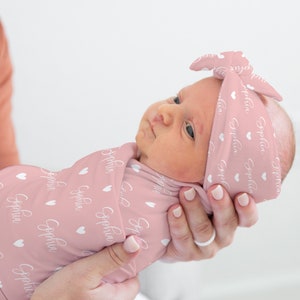 May include: A pink swaddle blanket with white hearts and the name "Sophia" printed on it. A baby is wrapped in the blanket and wearing a matching pink headband with the same pattern.